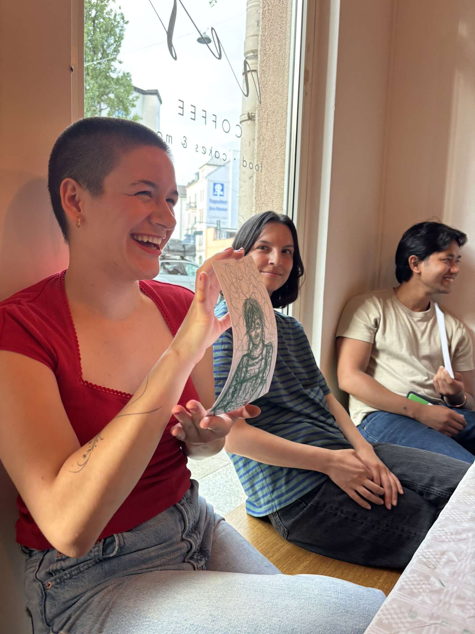 Laughing participant in a red top holding up a green pen portrait while two others smile beside her at a cafe window